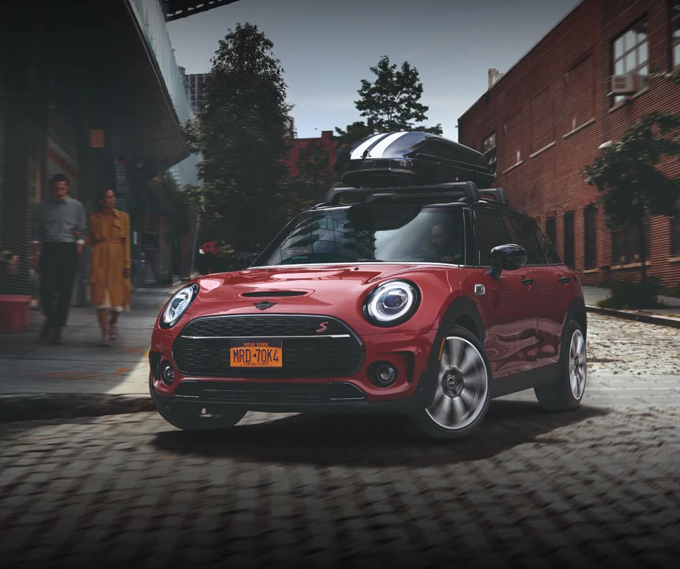 Front three-quarter view of a 2026 MINI Cooper S 4 Door in Chili Red II with a roof cargo box driving on a cobblestone street in an urban city setting.