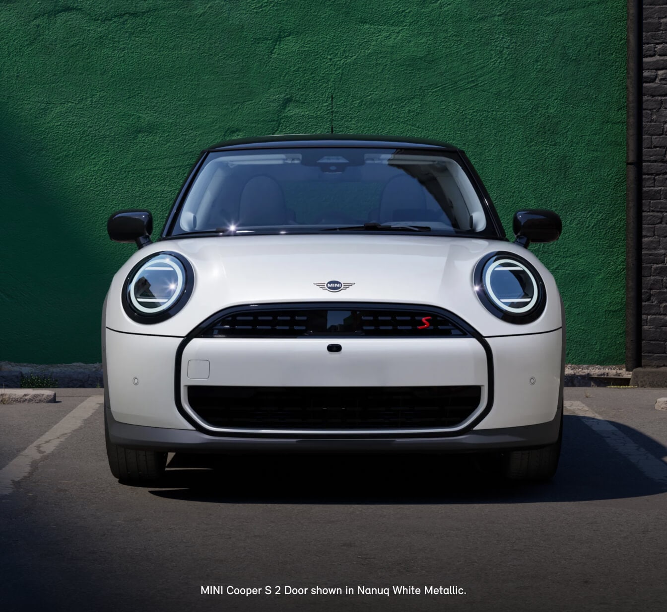 Front view of a 2026 MINI Cooper S 2-Door in Nanuq White Metallic with a black roof parked in front of a textured green wall.