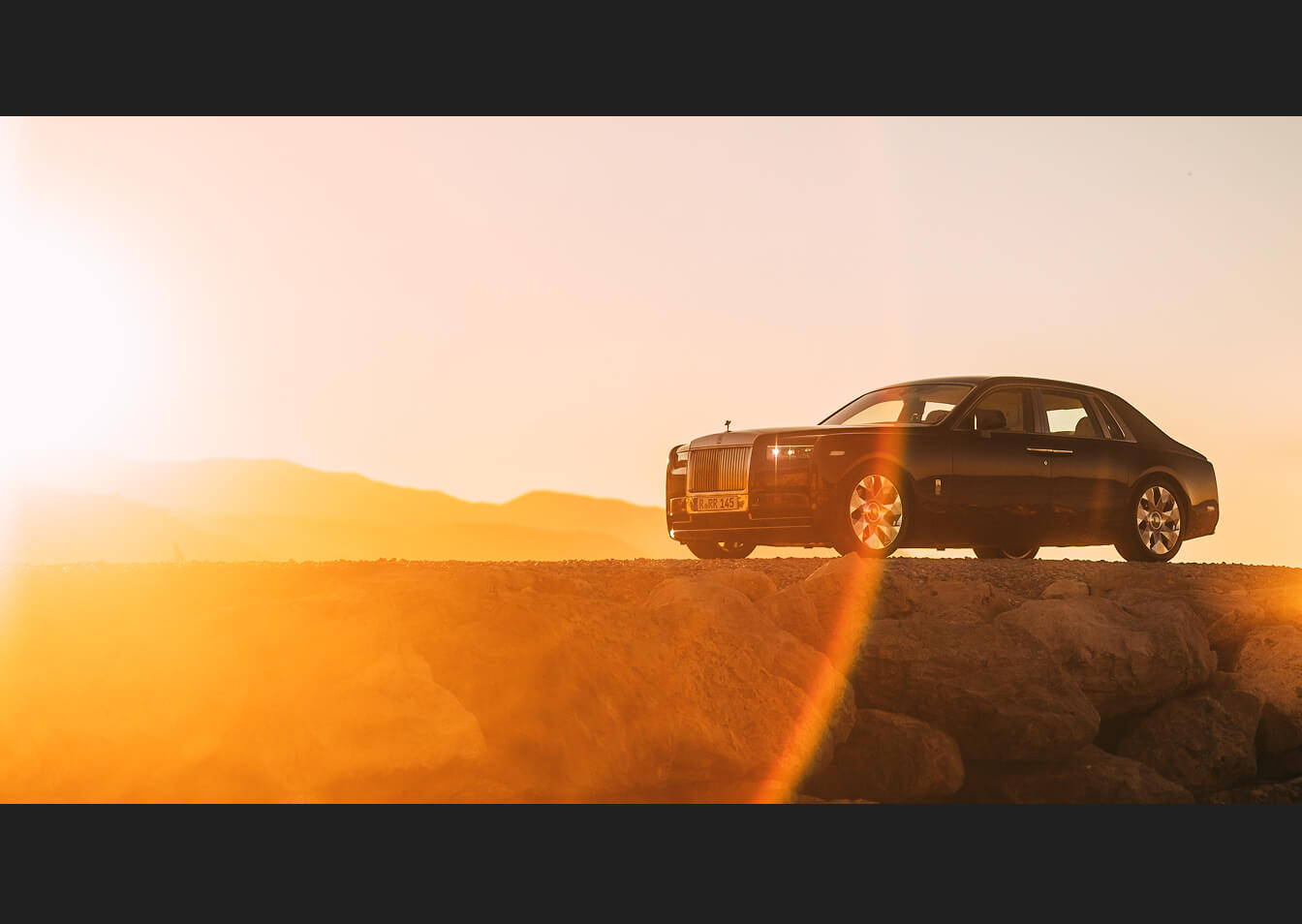 Side three-quarter view of a 2027 Rolls-Royce Phantom Series II finished in dark paint, parked on a rocky elevated ridge against a warm golden sunset.