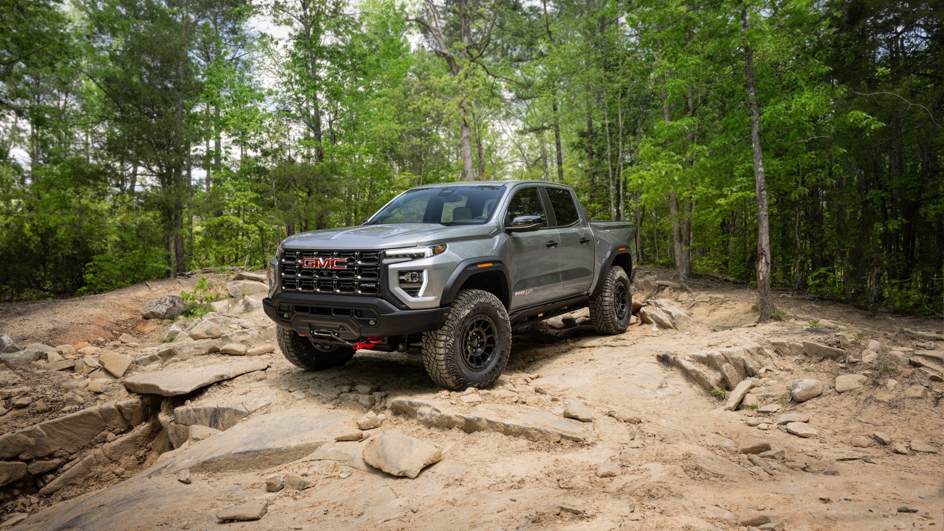 Gray GMC Canyon AT4X Crew Cab with a front winch climbing a rocky trail in a green forest
