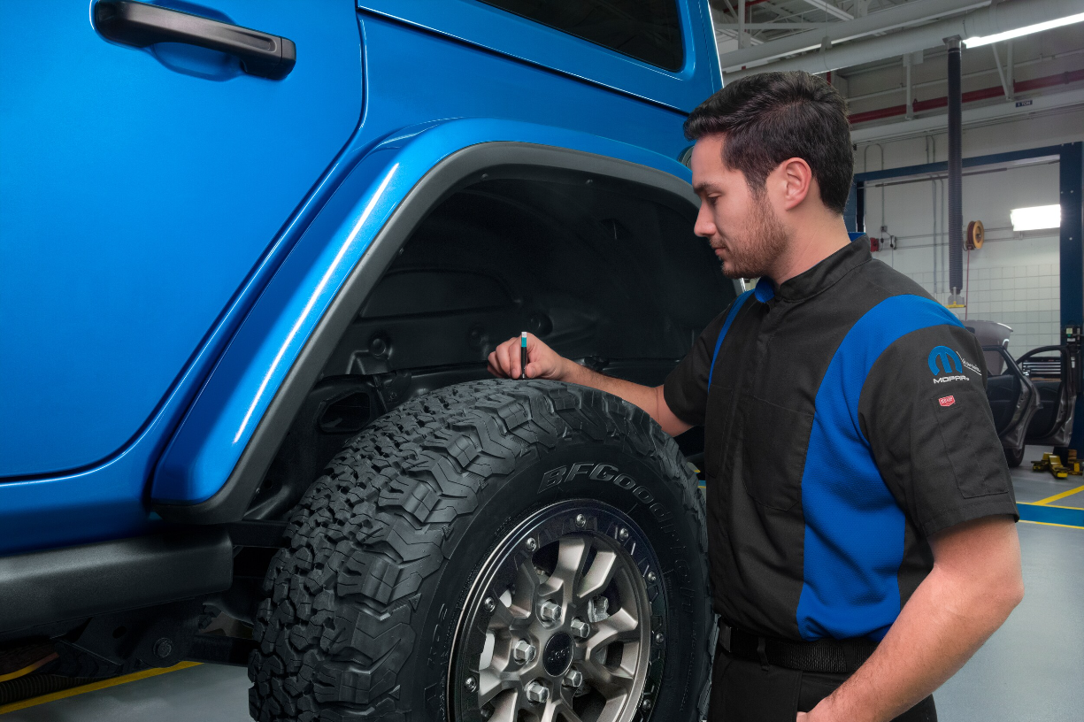 Technician checking tread depth on an all-terrain tire of a blue Jeep Wrangler in a service bay