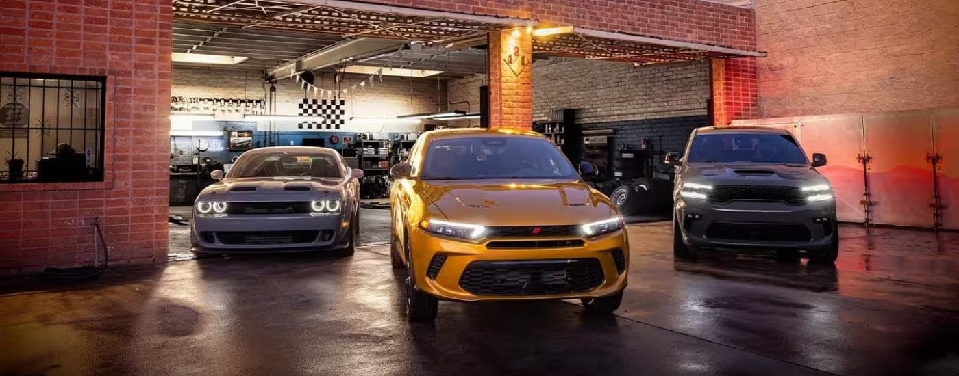 A yellow 2023 Dodge Hornet, a grey Durango, and a silver Challenger parked near garage.