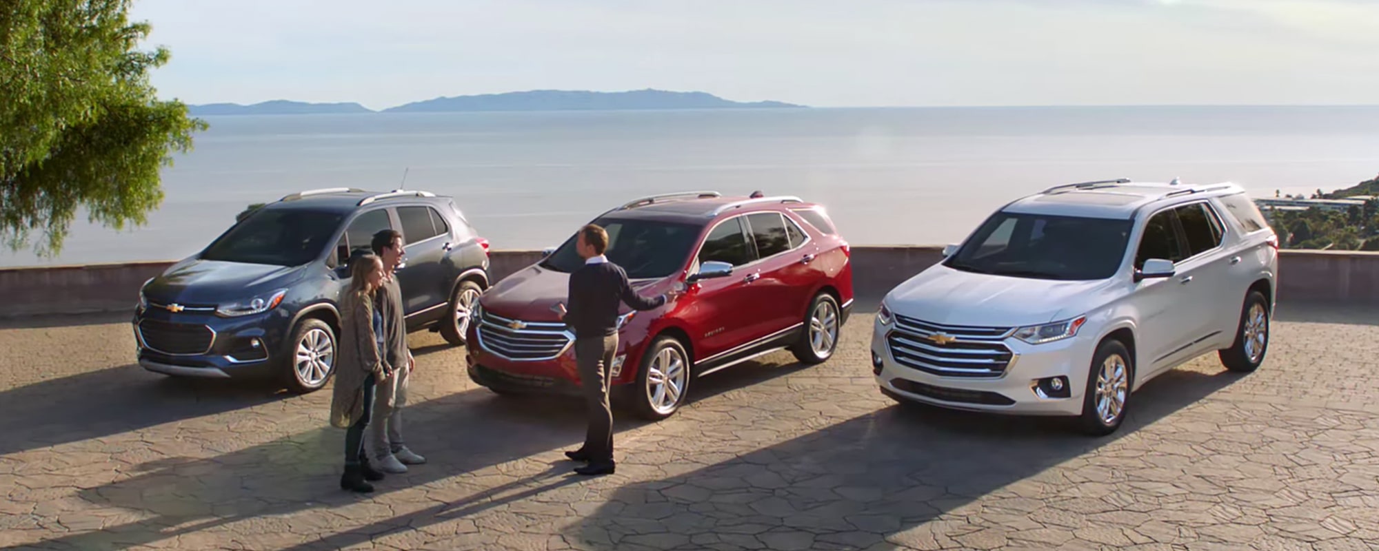 A man showing off a trio of gray, red, and white Chevy SUVs to a couple