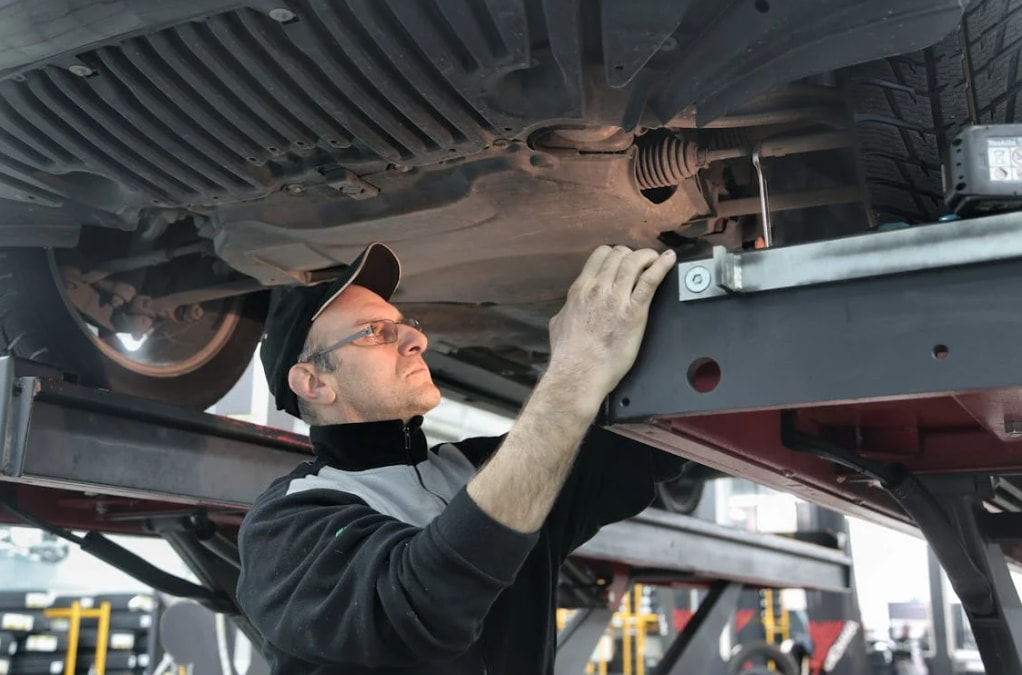Man in baseball cap and black jacket standing under a vehicle raised on a mechanic's lift