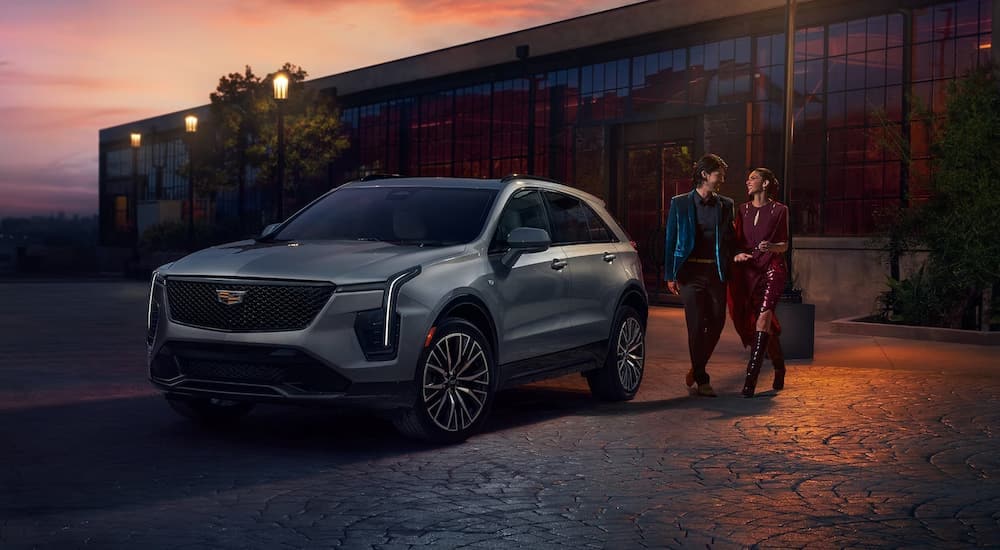 A silver 2024 Cadillac XT4 is shown parked outside of a large building during a sunset.