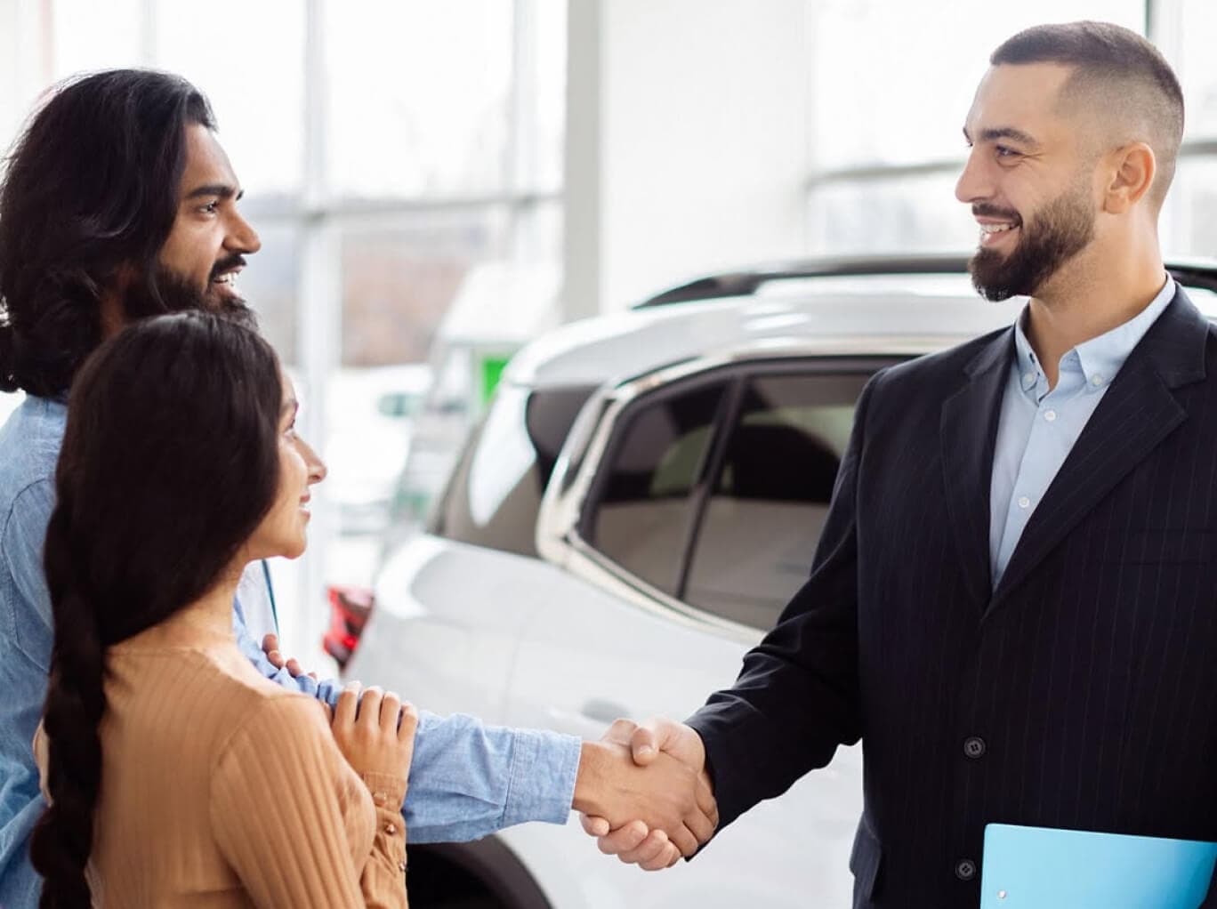 A sales associate in a suit shakes hands with a smiling couple in a bright, clean dealership.