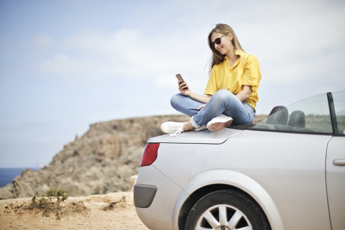 Woman in yellow shirt sitting on the back of a convertible car looking at her smartphone