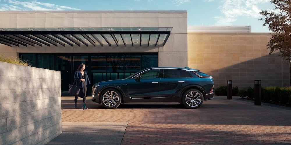A woman walking toward a dark blue 2026 Cadillac Lyriq SUV parked in front of a modern building