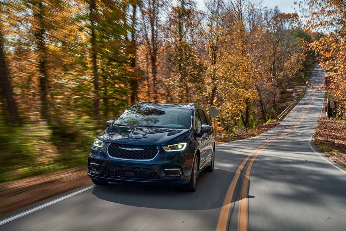 A navy blue 2022 Chrysler Pacifica Hybrid driving on a road at the edge of a forest in the fall season