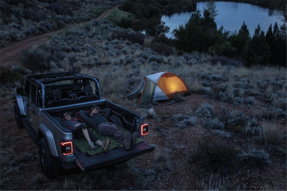 un couple en camping couché dans la boite d'un Jeep Gladiator près d'une tente sur le bord d'un lac le soir