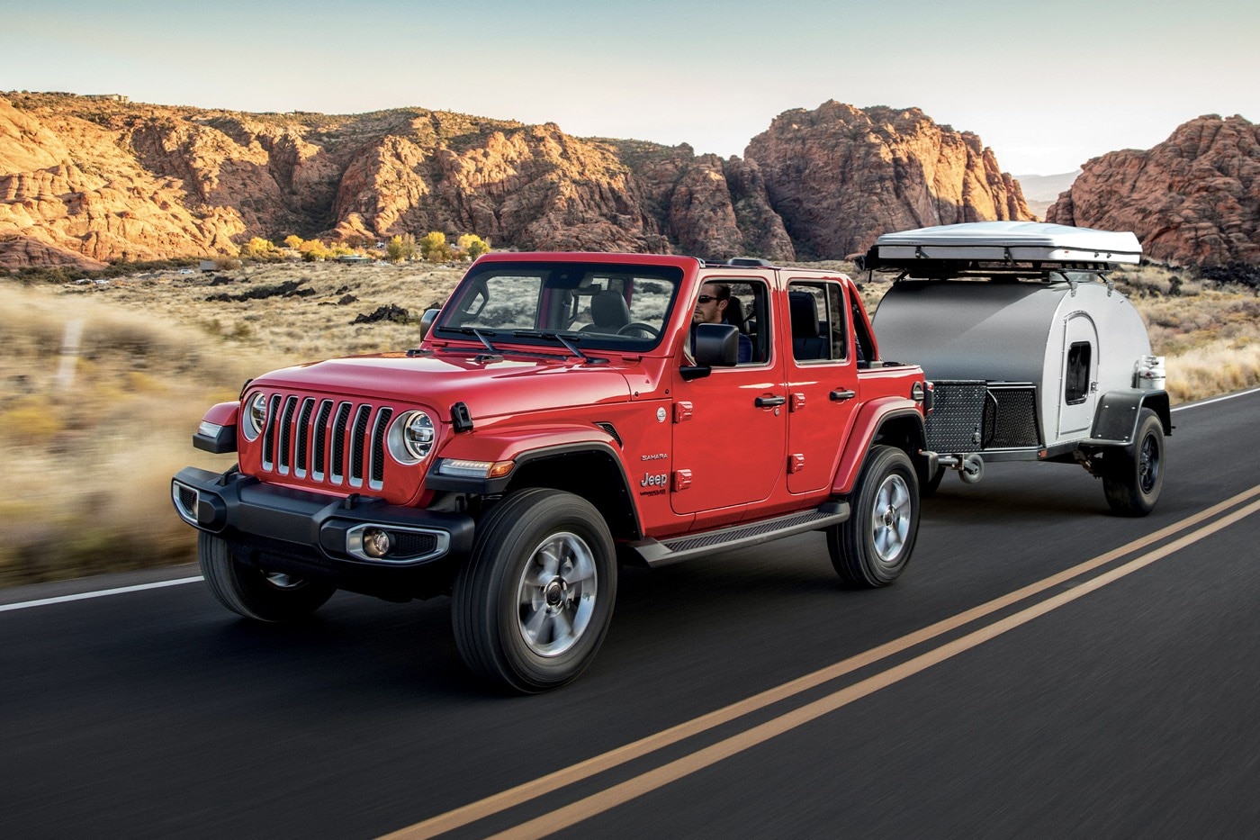 Un Jeep Wrangler Unlimited Sahara 2022 rouge tirant une petite autocaravane sur fond de désert