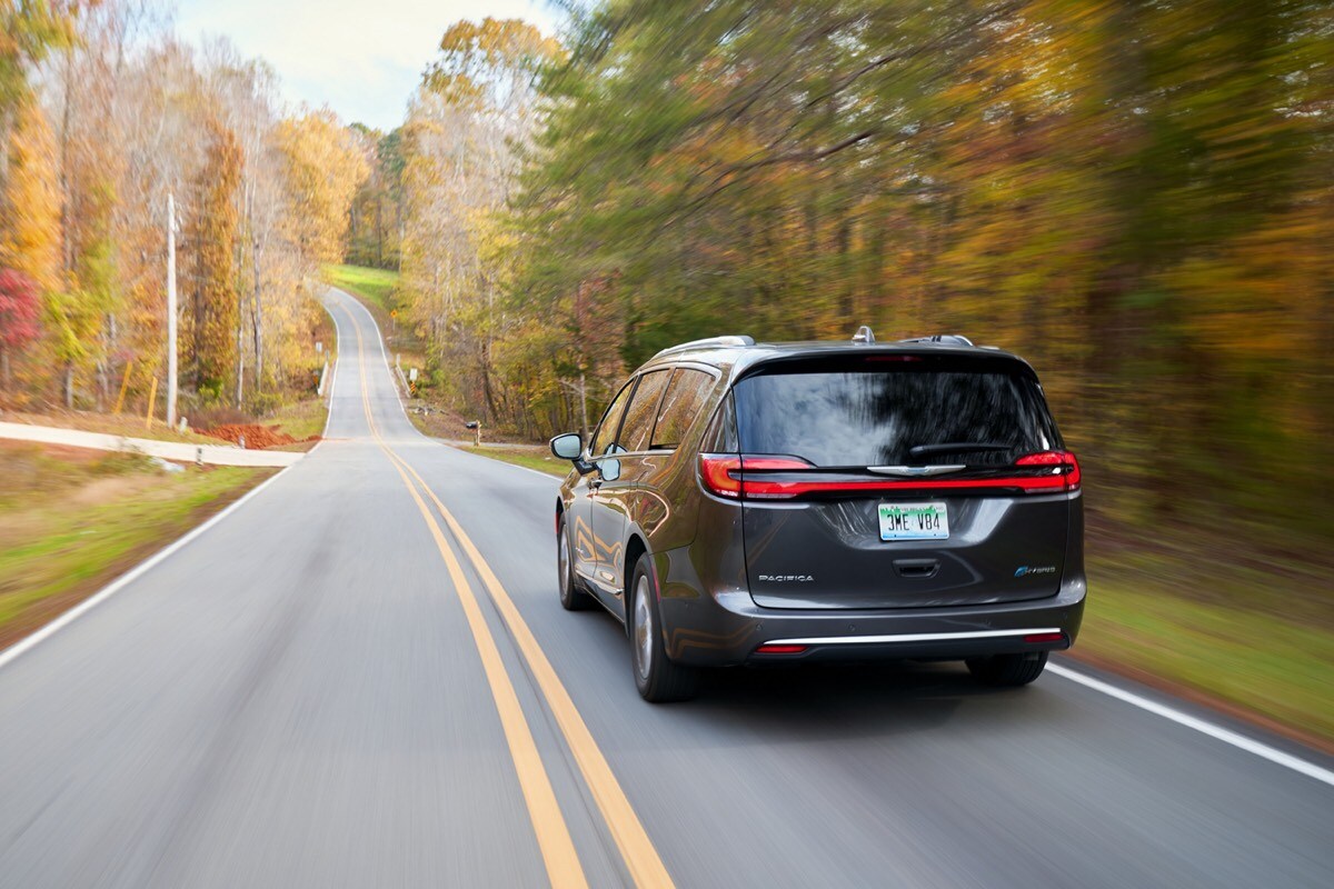 Rear 3/4 view of a 2022 Chrysler Pacifica Hybrid driving on a road at the edge of a forest