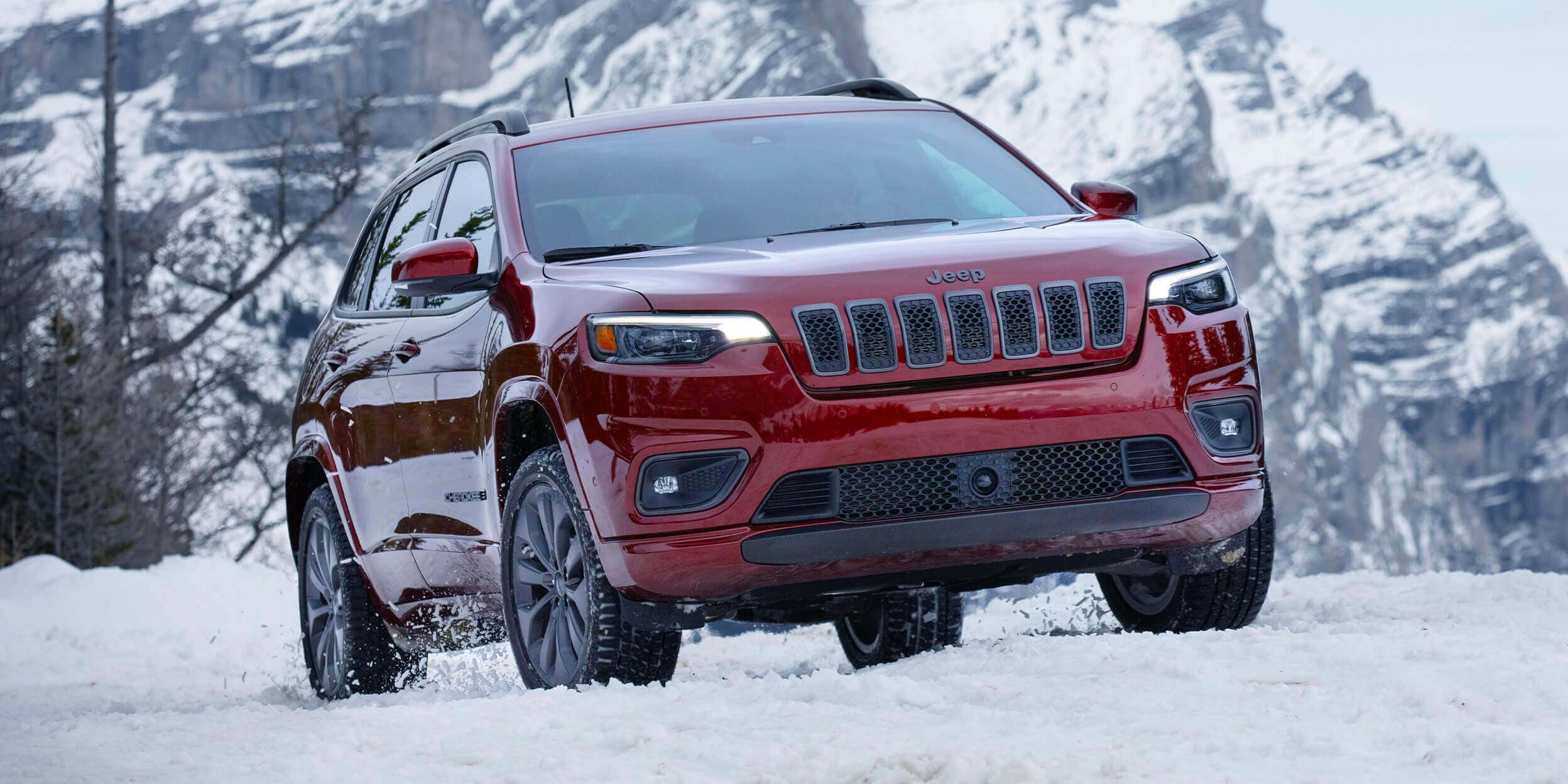 A velvet red 2022 Jeep Cherokee driving through the snow with the mountains visible in the distance