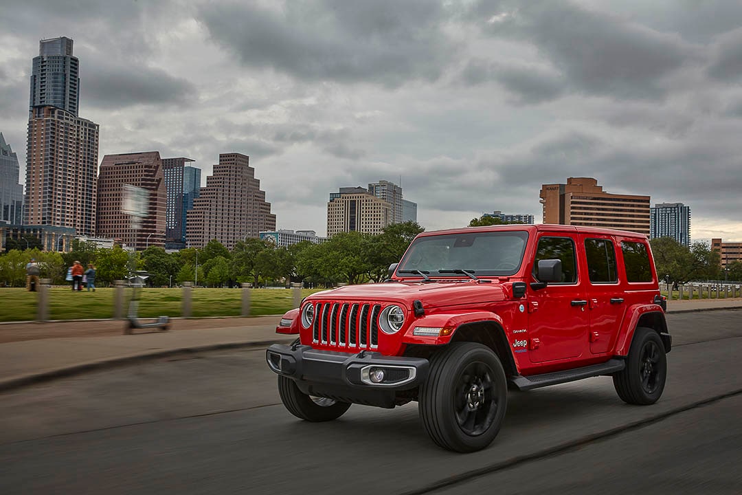Front 3/4 view of a red Jeep Wrangler Sahara 4xe driving on a road not far from a big city