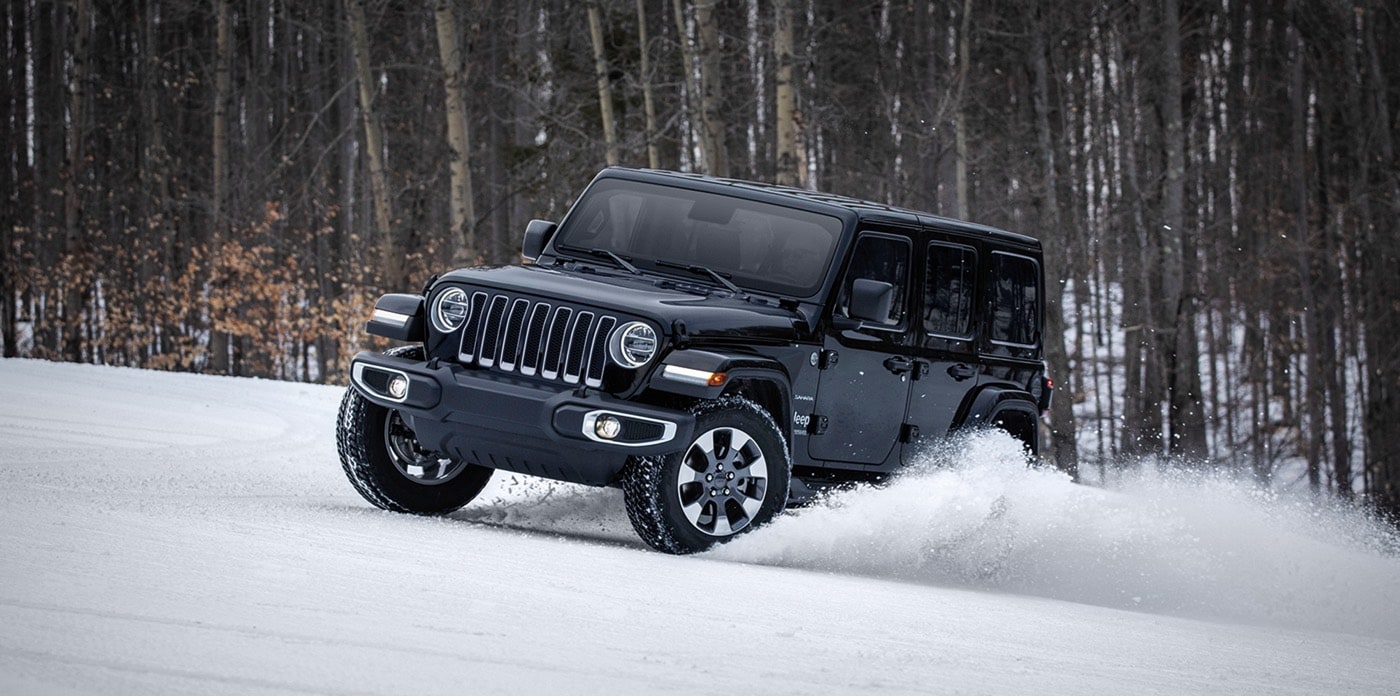 2022 Jeep Wrangler Wrangler Unlimited Sahara in black driving near forest, lifting snow as it moves