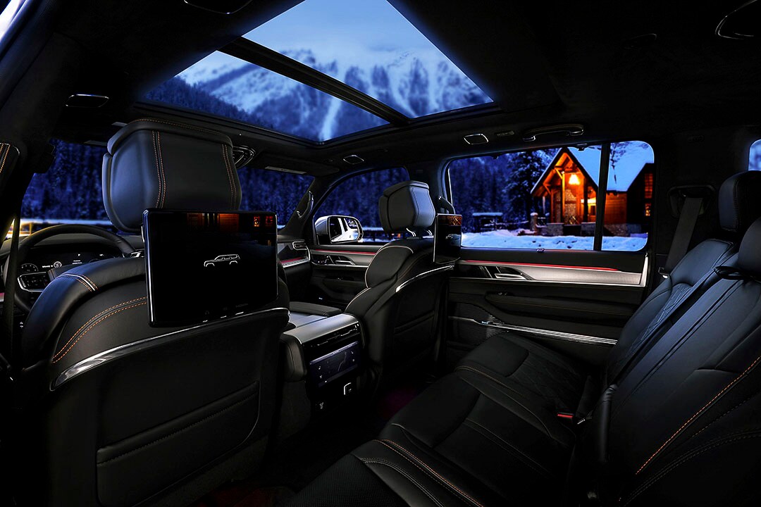 Vue intérieur d'un Jeep Wagonneer en cuir noir avec lignes rouges à partir de la seconde rangée vers le passager avant, avec toit ouvrant, grande fenêtre arrière, 2 écrans plats et une console, tout cela sur fond d'un chalet enneigé en montagne, au crépuscule