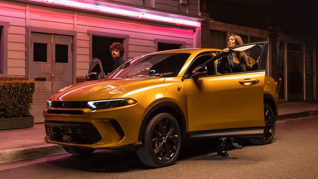 An adult woman and adult man stepping out of a parked yellow Dodge Hornet