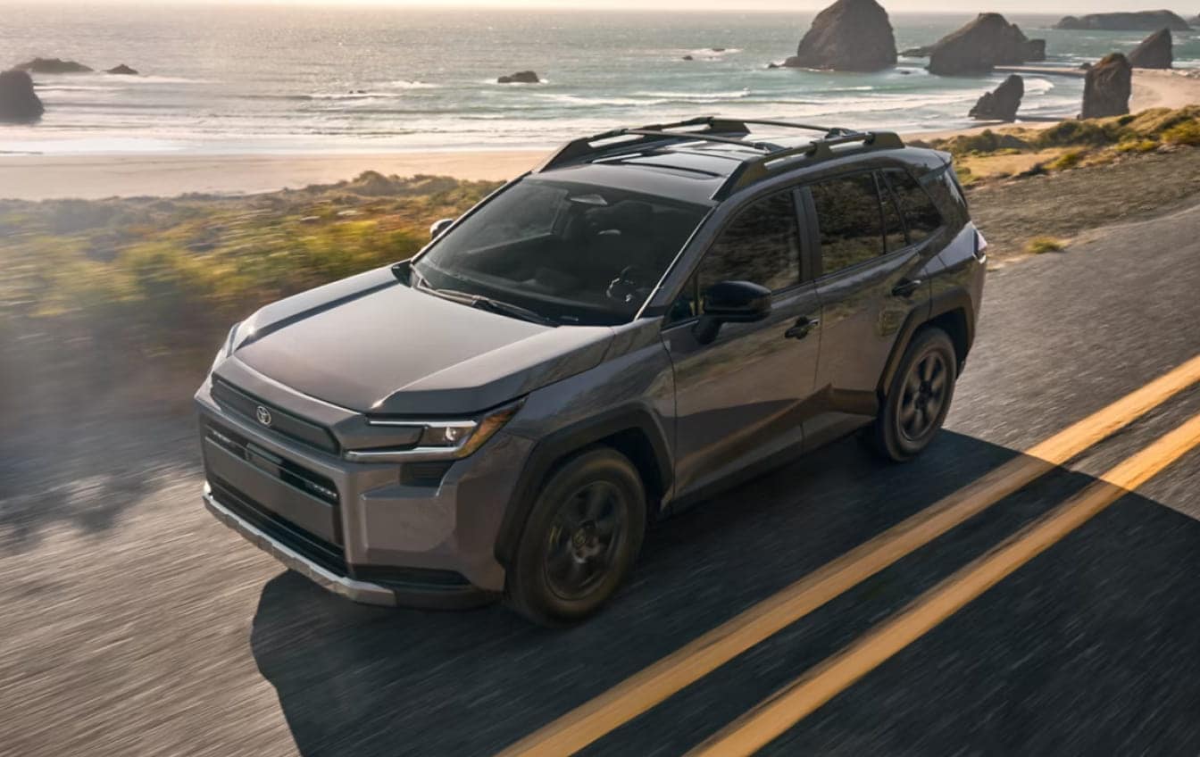 2026 RAV4 Woodland in Urban Rock driving along a scenic coastal road with rugged cliffs and ocean waves in the background.