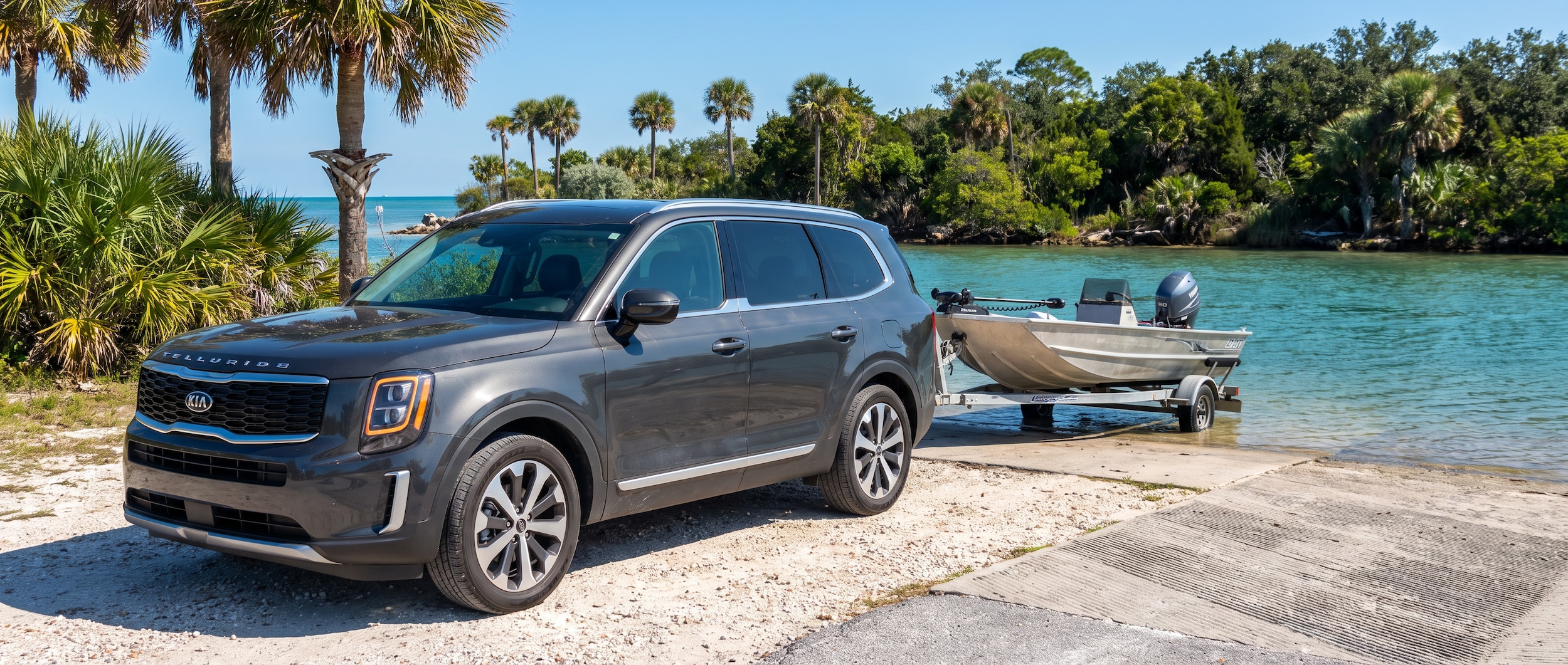 A used Kia Telluride parked near a Gulf Coast boat ramp on a bright summer day in Panama City, Florida, with calm turquoise water and a small aluminum fishing boat on a trailer visible in the background, lush subtropical greenery framing the scene.