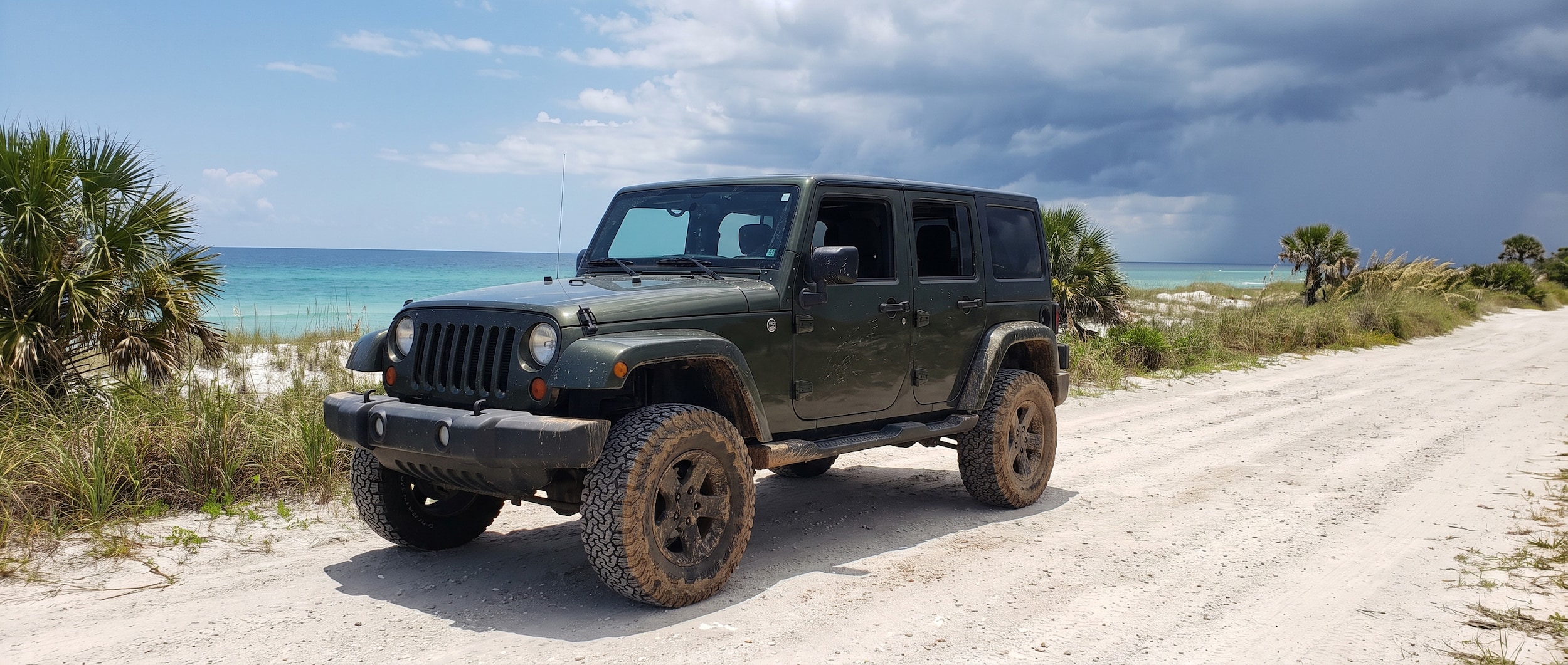 A used dark-colored Jeep Wrangler 4-door parked on a sandy Florida Panhandle coastal road in summer, rugged all-terrain tires and high ground clearance visible, blue Gulf sky and afternoon storm clouds in the background