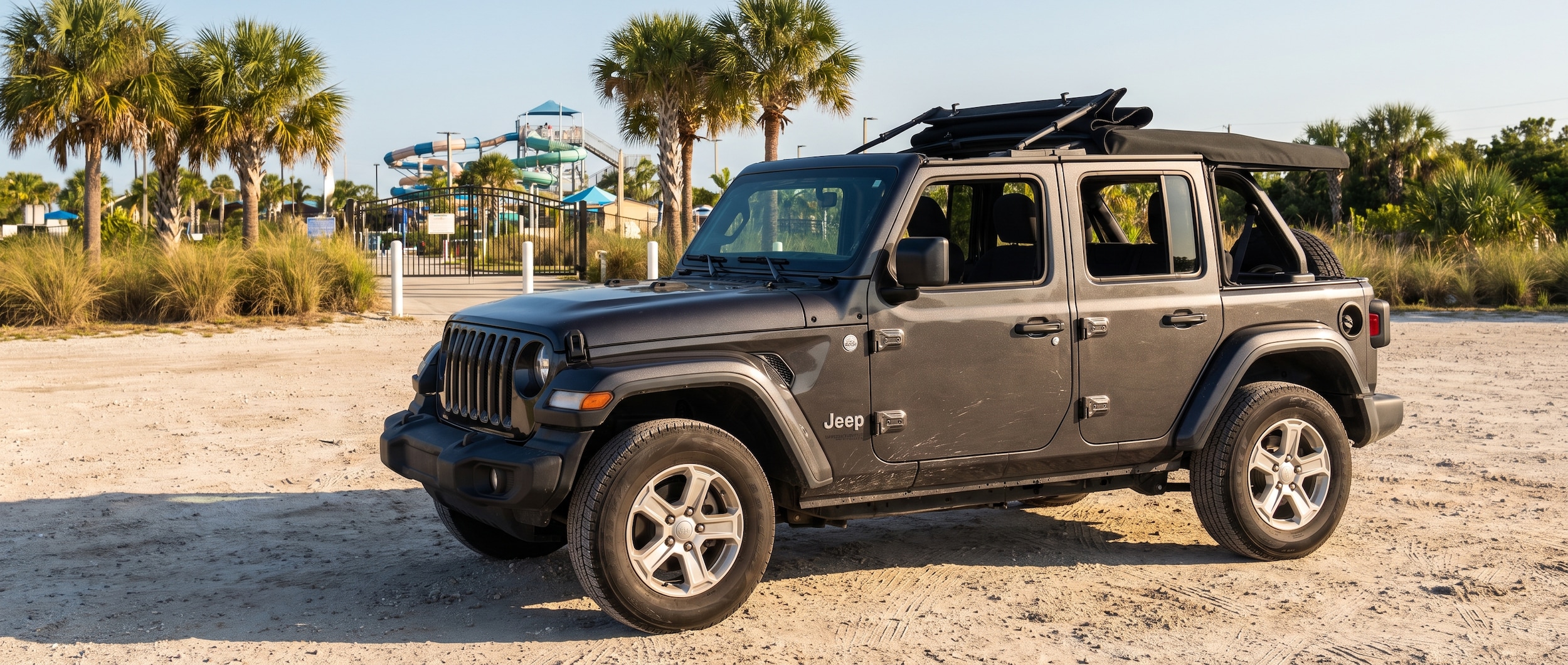 A used Jeep Wrangler JL (2022 or 2023, dark green or granite crystal) parked at a sandy Gulf Coast parking area near a waterpark entrance, Florida summer light, palm trees in background, open soft top