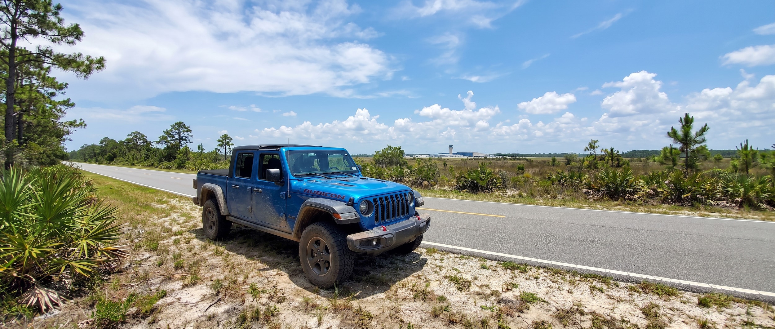 A used Jeep Gladiator in Hydro Blue or Granite Crystal metallic parked on West Bay Parkway near Northwest Florida Beaches International Airport with the soft top removed, Gulf Coast summer sky overhead, 2022 or 2023 model year