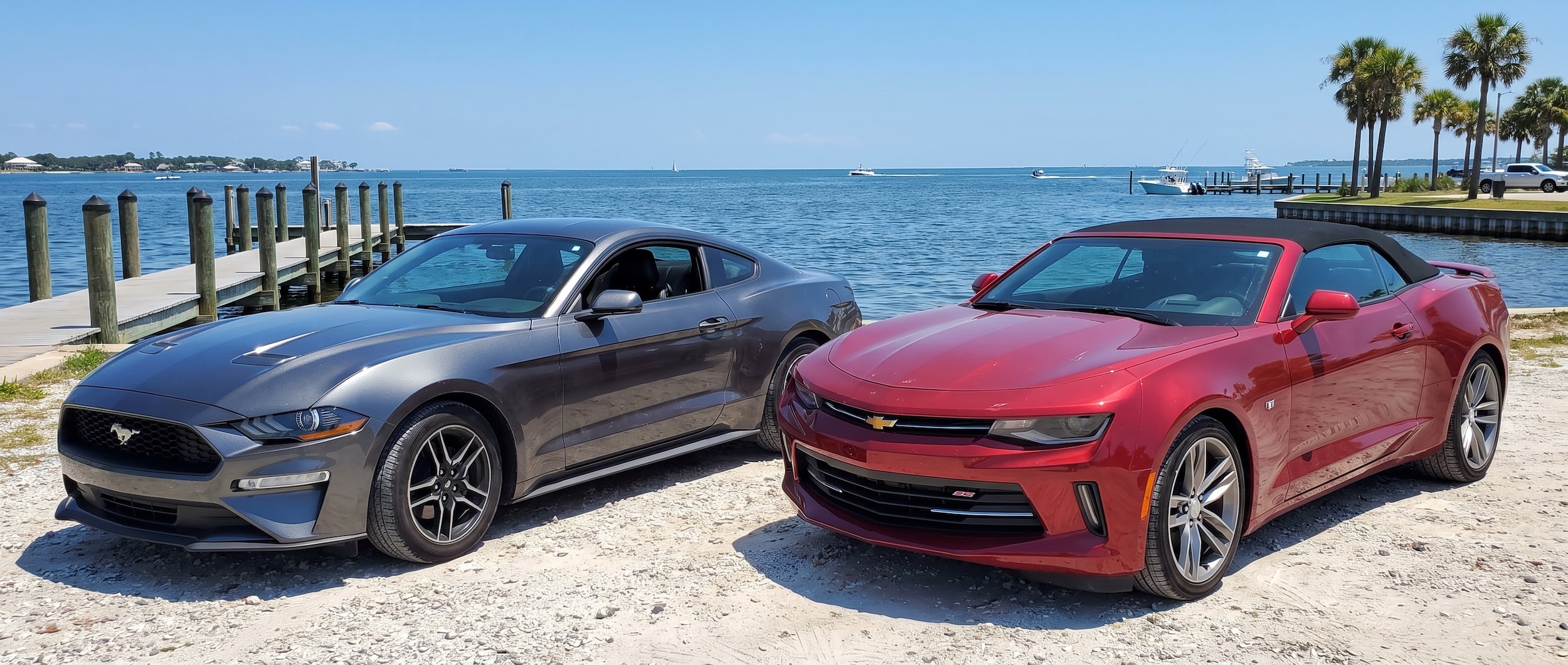 A used Ford Mustang and a used Chevrolet Camaro parked side by side on a sunlit waterfront lot near St. Andrews Bay in Panama City, Florida, with blue water visible in the background on a bright summer day