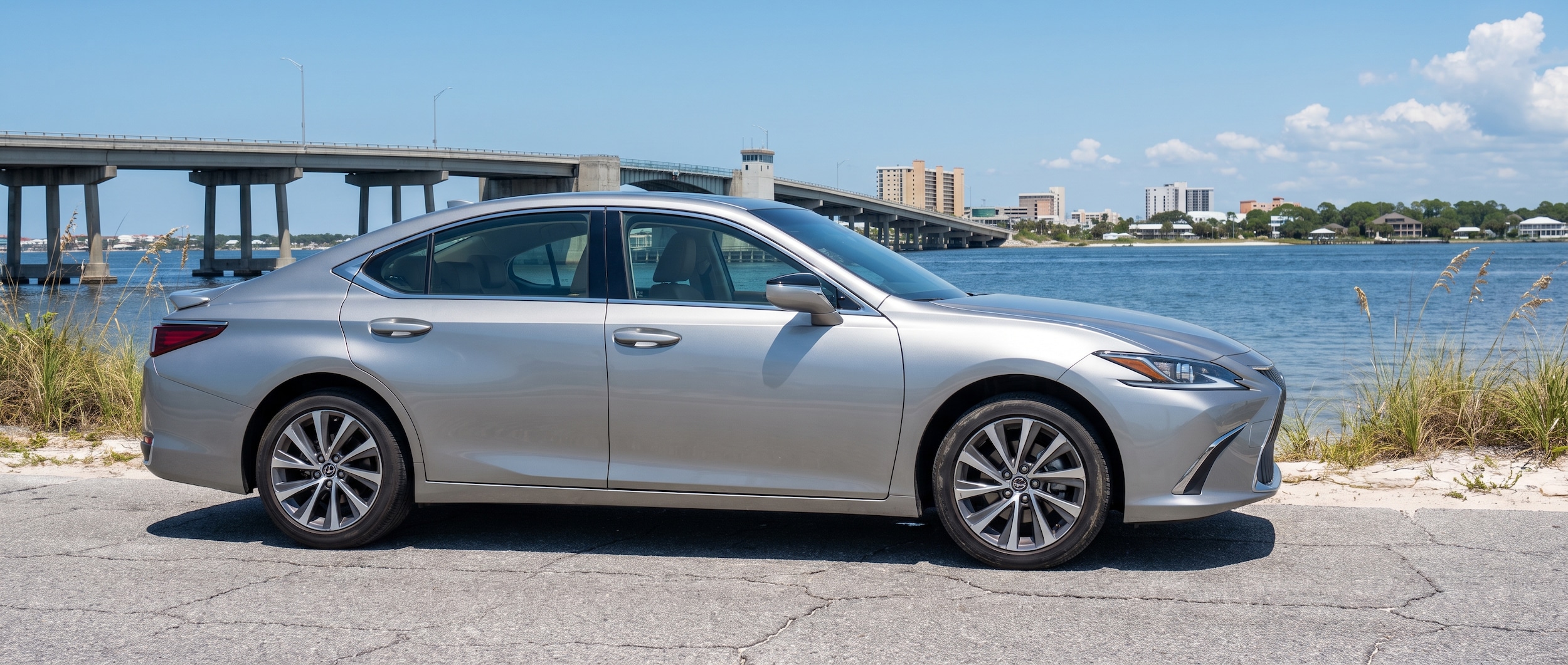 A refined used luxury sedan -- a silver Lexus ES 350 -- parked on a Gulf Coast road with the Hathaway Bridge and Panama City skyline visible in the background under a bright summer sky, 2022 model year