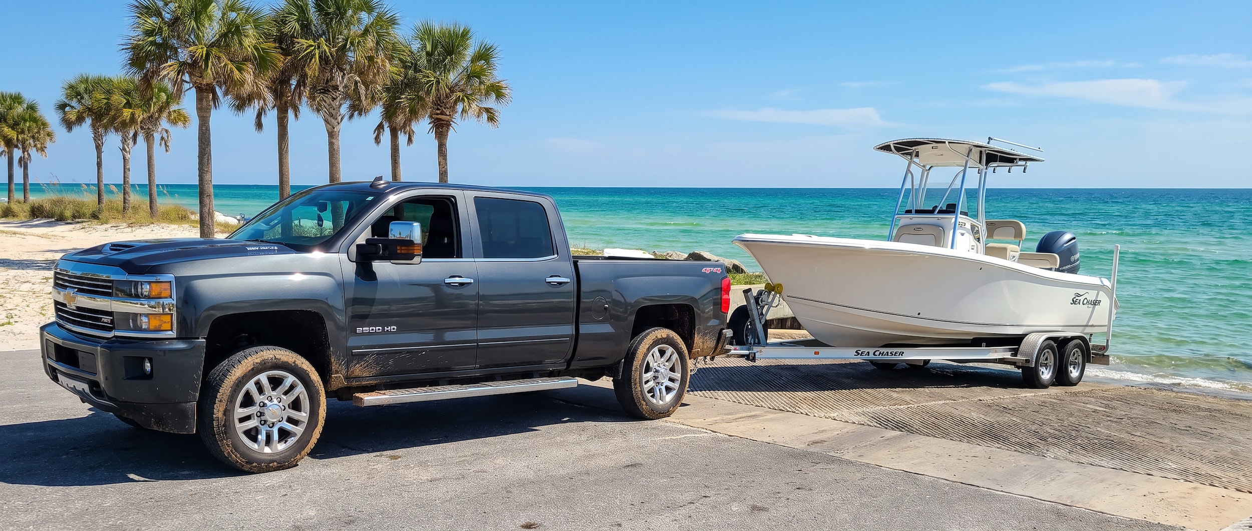 A used Chevrolet Silverado 2500 HD in dark metallic gray backing a bay boat trailer down a concrete boat ramp on a bright Florida summer day, palm trees and emerald Gulf water visible in the background, Panama City Beach area