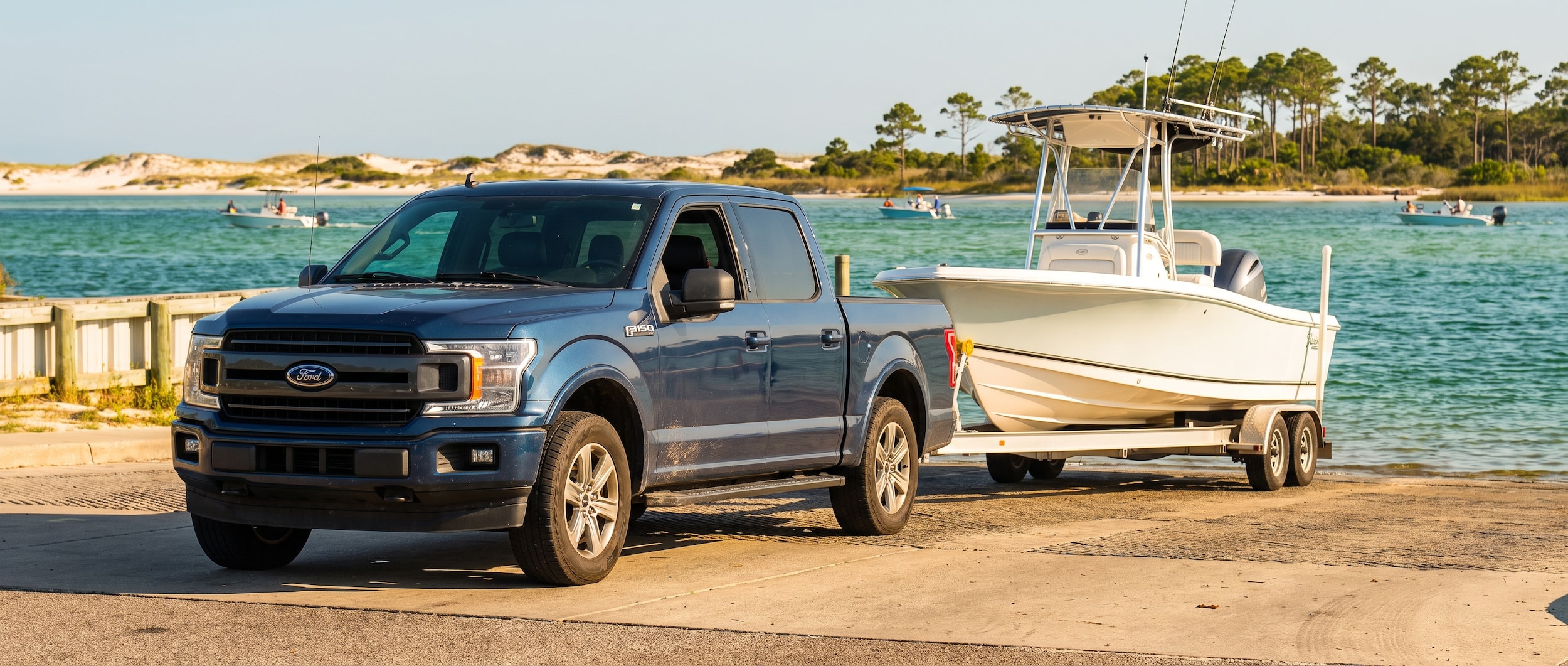 A used full-size pickup truck backed up to a sunny boat ramp on Grand Lagoon at St. Andrews State Park near Panama City, Florida, with a center-console fishing boat on a trailer, emerald-green water visible in the background, summer morning light