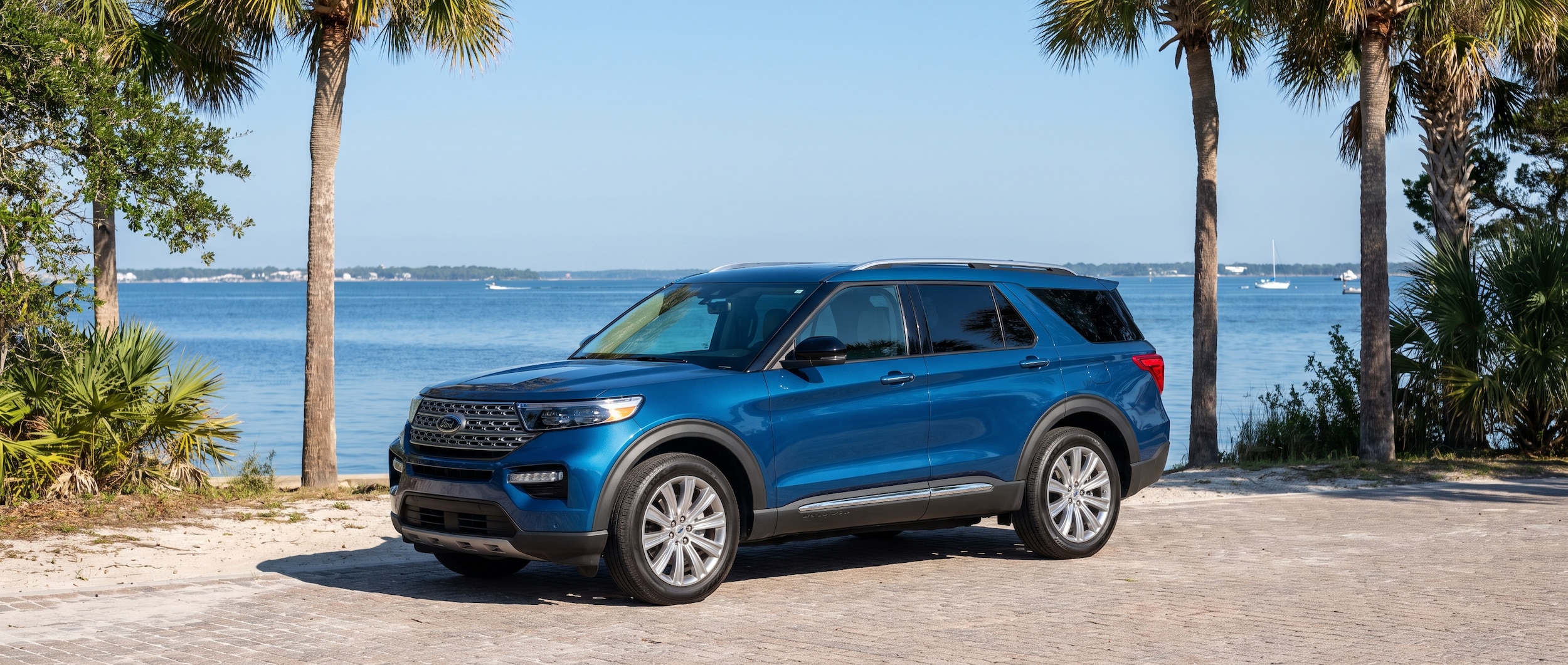 A 2026 Ford Explorer in a deep blue or silver finish parked near the St. Andrews Bay waterfront in Panama City, FL on a bright summer morning, with the bay visible in the background and Florida palms framing the scene