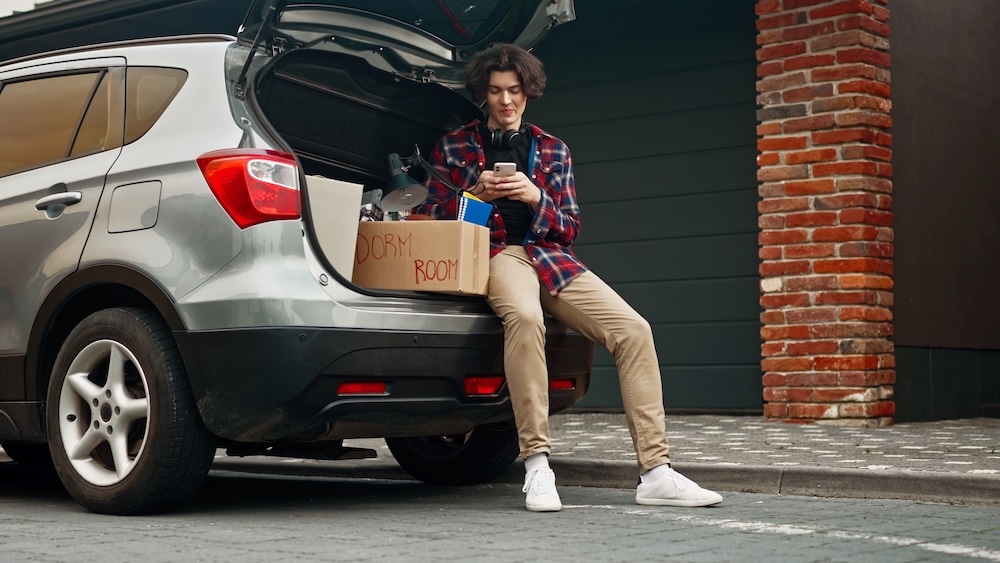 A teenager sitting in the open trunk of a silver SUV, beside boxes marked “Dorm Room”