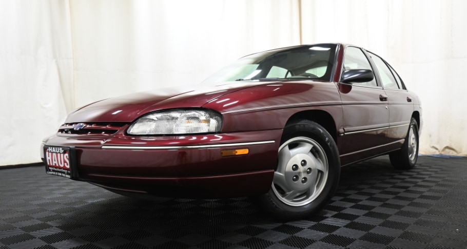 Burgandy 1998 Chevrolet Lumina LS in photo booth at Haus Auto Group with a driver side and front wheel turned all the way to the right has a white wall curtain behind and the car is on a black plastic square patterned floor
