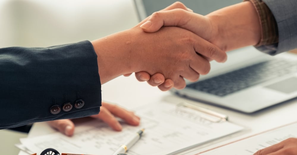 The hands of two white people in blazers clasped in a handshake, with a laptop and paperwork out of focus in the background