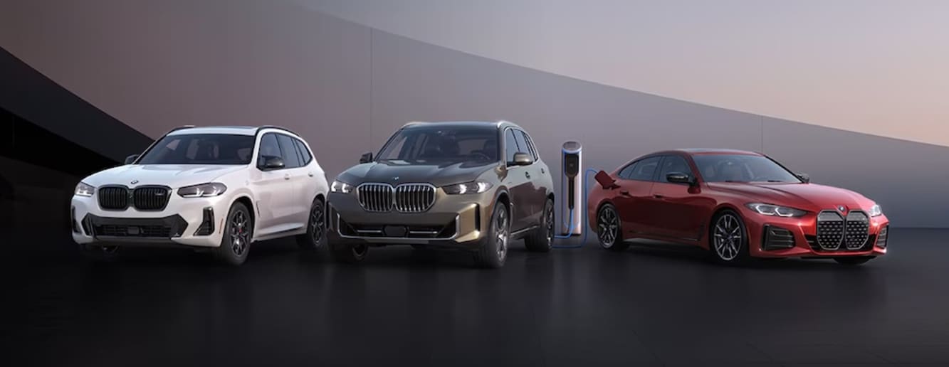 The BMW vehicles, one white, one gray, and one red, parked in front of a sloping gray wall, the two rightmost vehicles plugged into a charging station