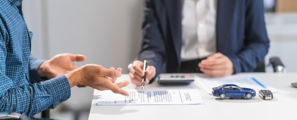 Two people discussing paperwork at a table, with a calculator, toy car, and car key between them 