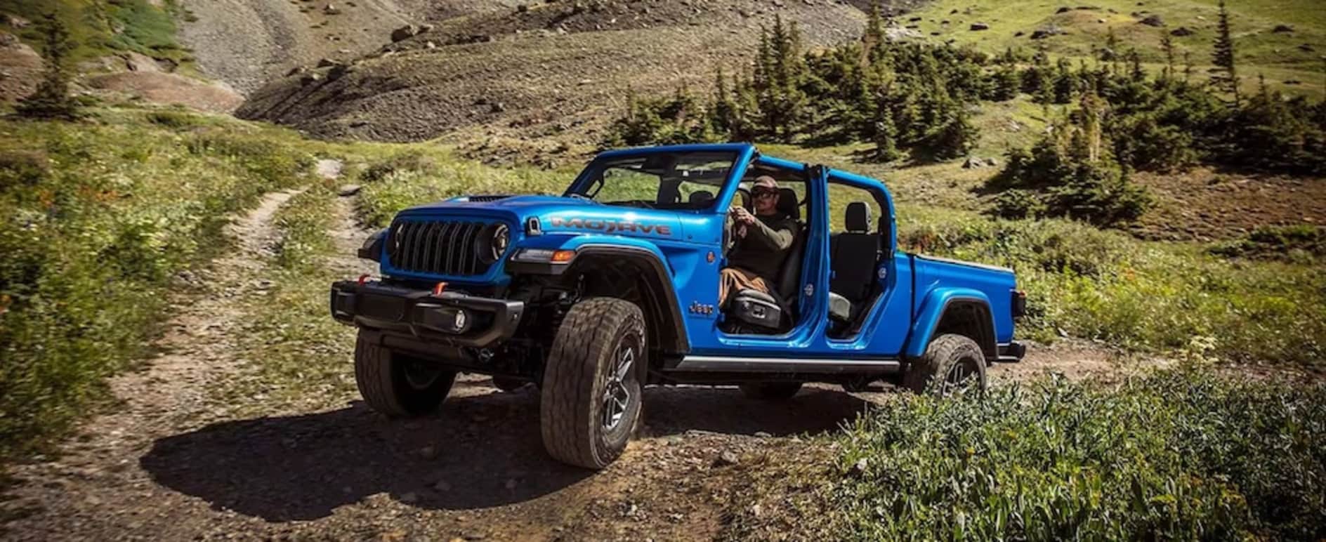 A white man in a baseball cap driving a bright blue 2026 Jeep Gladiator up a grassy hill, with rocky mountains and pine trees in the background