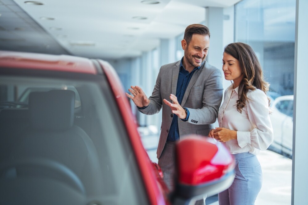 A man and a woman with light skin and brown hair standing in a car dealership, discussing a red vehicle