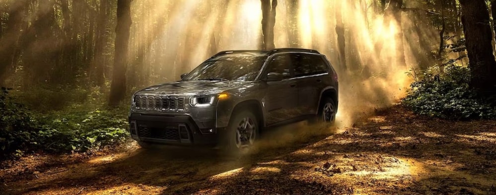 A gray 2026 Jeep Cherokee SUV driving down a dirt road through trees, with golden sunlight shining down