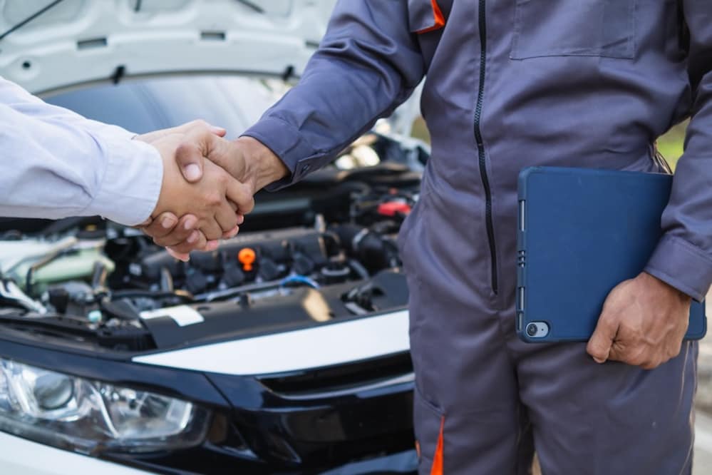 A person in coveralls holding a clipboard and shaking hands with a person in shirtsleeves, with a vehicle's engine in the background