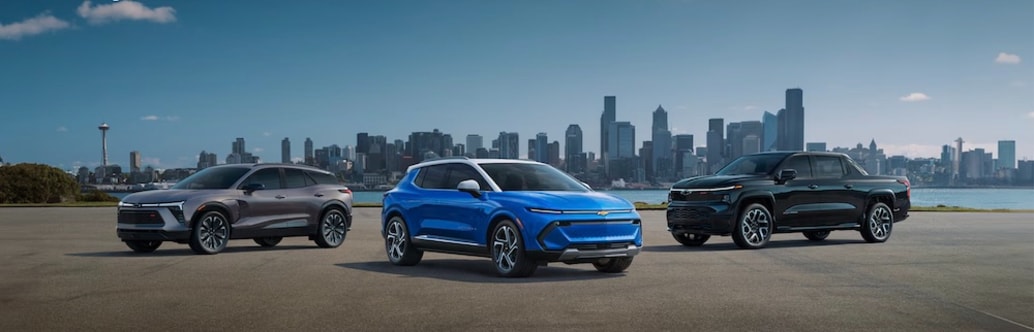 3 Chevrolet vehicles, a gray SUV, a blue SUV, and a black truck, parked by a river, with a cityscape in the distance