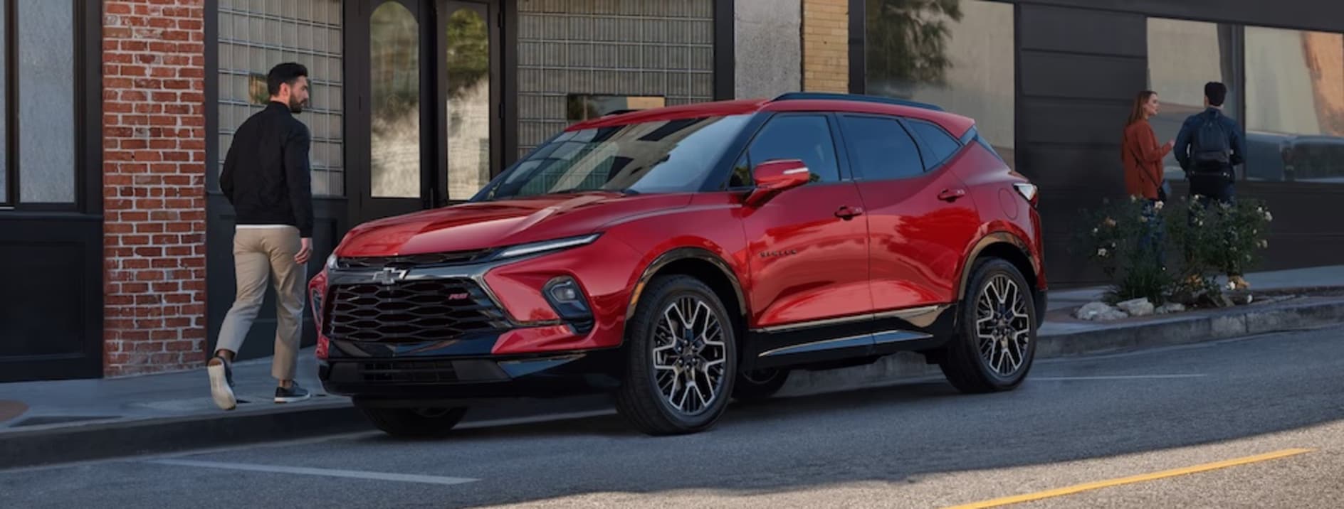 A man in a black sweater walking past a red 2026 Chevrolet Blazer SUV parked on a city street