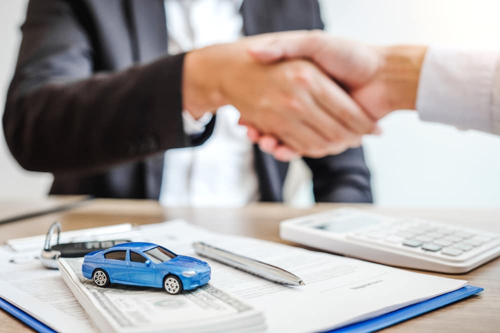 Two people shaking hands across a desk, with a toy car, paperwork, cash, and a calculator between them