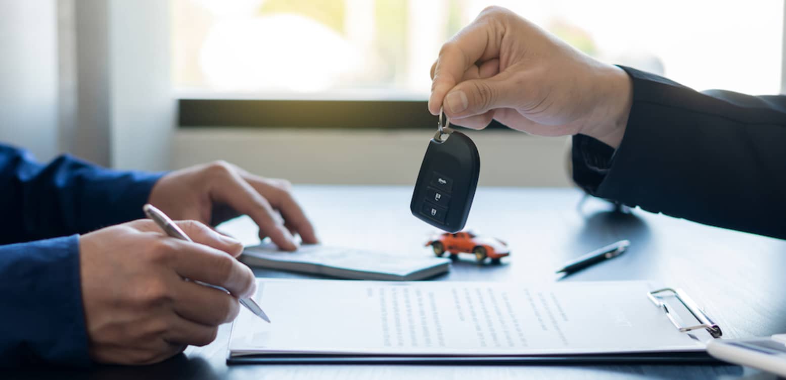 A closeup of two white people's hands on a desk, one holding a pen to sign paperwork, the other holding out a car key