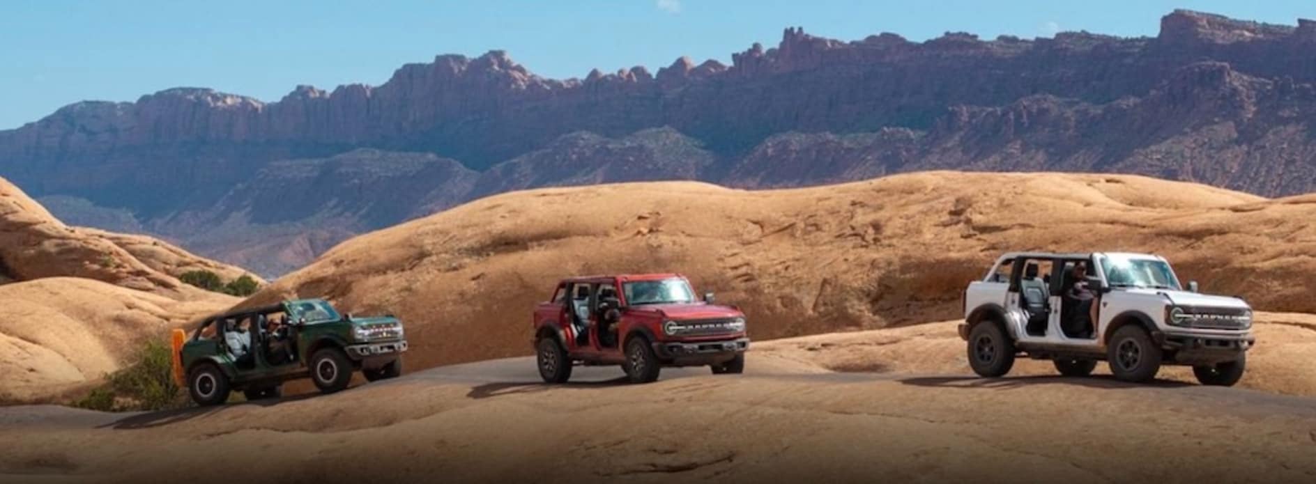 AThree Ford Broncos, one green, one red, one white, driving across bare dirt hills with mountains in the background