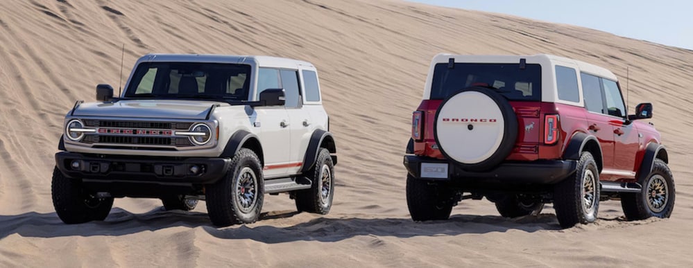 Two 2026 Ford Bronco SUVs, one white and one red, parked in opposite directions on a sand dune