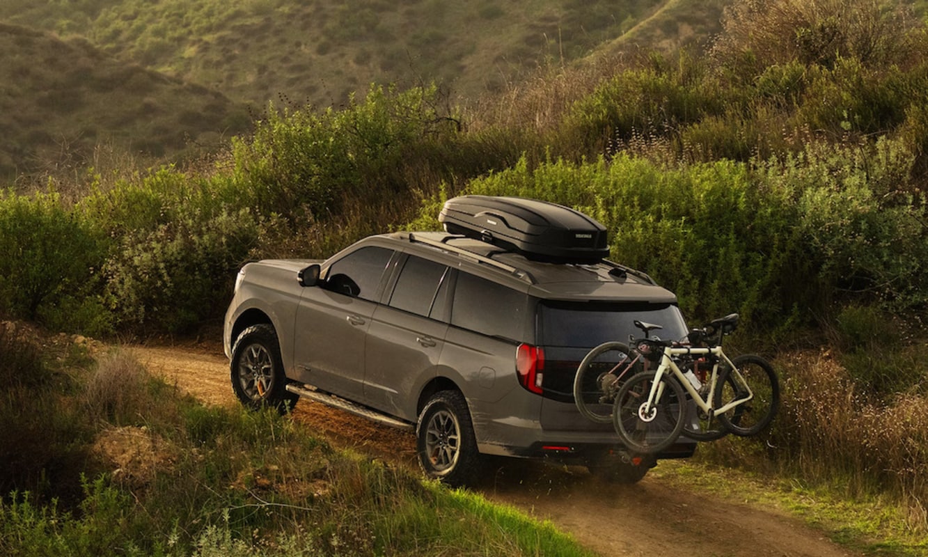 An olive green 2026 Ford Expedition SUV drives over a dirt lane through hilly terrain