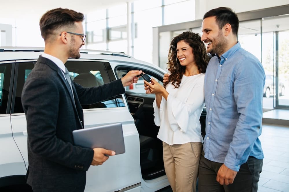A car salesman hands a couple a car key inside a car dealership
