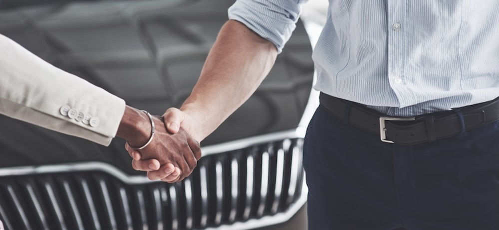 Two people shaking hands in front of the hood of a car