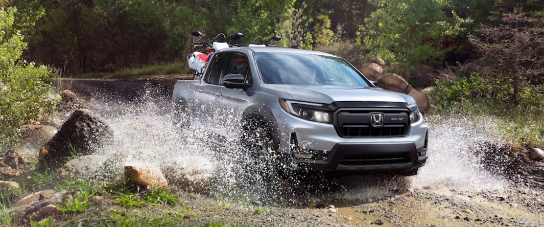 A silver 2026 Honda Ridgeline pickup truck driving out of a riverbed, splashing up water from its wheels